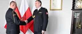 Strażak w mundurze wyjściowym ze sznurem z czerwoną teczką trzyma dłoń drugiego strażaka w mundurze wyjściowym ze sznurem za nimi są trzy flagi Polski na ścianie wisi obraz obok stoi zegar a w ścianie są wyłączniki. 