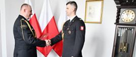 Strażak w mundurze wyjściowym ze sznurem z czerwoną teczką trzyma dłoń drugiego strażaka w mundurze wyjściowym ze sznurem za nimi są trzy flagi Polski na ścianie wisi obraz obok stoi zegar a w ścianie są wyłączniki w rogu widać górę krzesła.