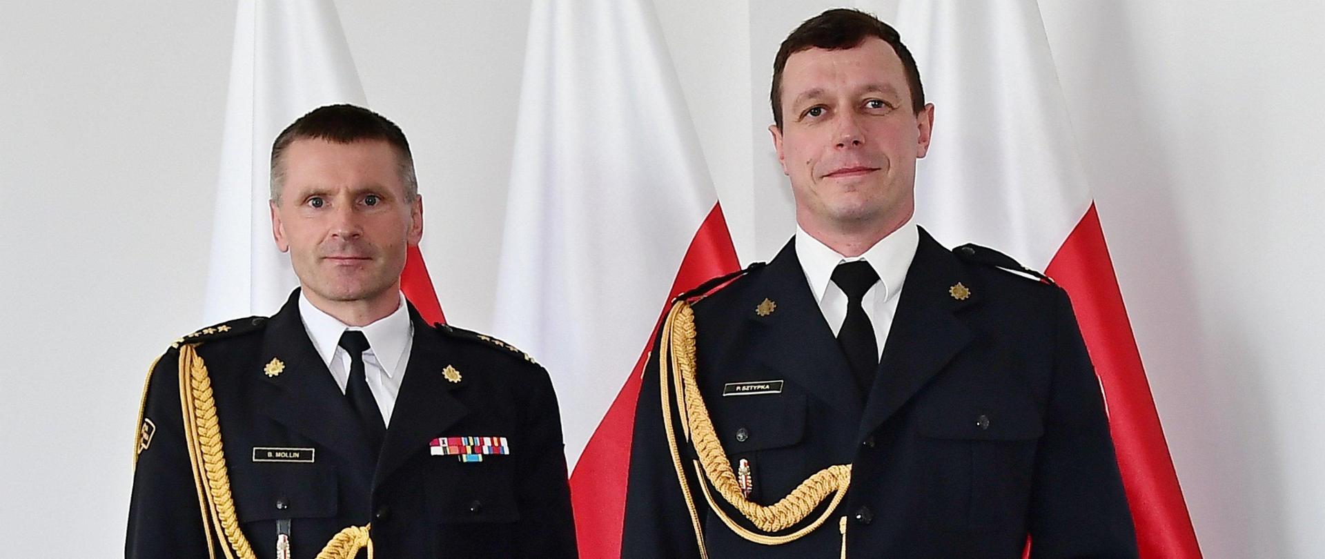 Dwóch strażaków w mundurach galowych stoi obok siebie jeden z nich trzyma czerwoną teczkę za nimi ustawione są trzy flagi Polski.