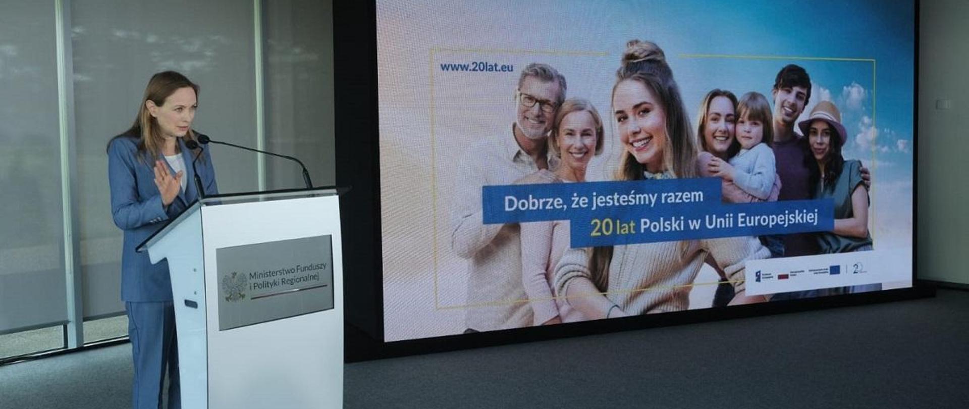 Minister funduszy i polityki regionalnej Katarzyna Pełczyńska-Nałęcz stoi przy mównicy, po jej lewej stronie na ekranie wyświetlone jest zdjęcie przedstawiające mężczyzn i kobiety, z dzieckiem i napisem "Dobrze, że jesteśmy razem. 20 lat Polski w Unii Europejskiej"
