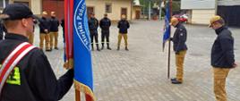 Zdjęcie zrobione na zewnątrz, na placu przed komendą podczas szkolenia członków pocztów sztandarowych. Strażacy stoją w szeregach, część z nich trzyma drzewce sztandarów.