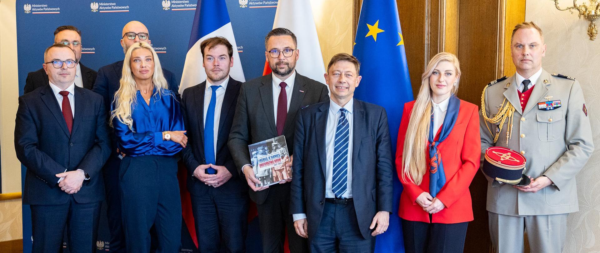 Członkowie delegacji stoją na tle flag Francji, Polski i Unii Europejskiej. Wiceminister Kulasek w środku. Na zdjęciu znajduje się 9 osób.