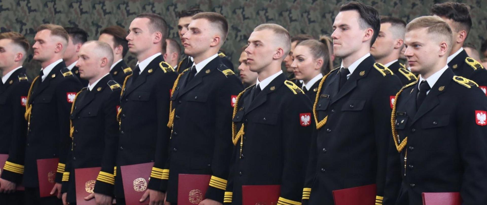 Podchorążowie Akademii Pożarniczej stoją w szeregu, trzymając w lewej dłoni przy boku dyplomy ukończenia studiów.