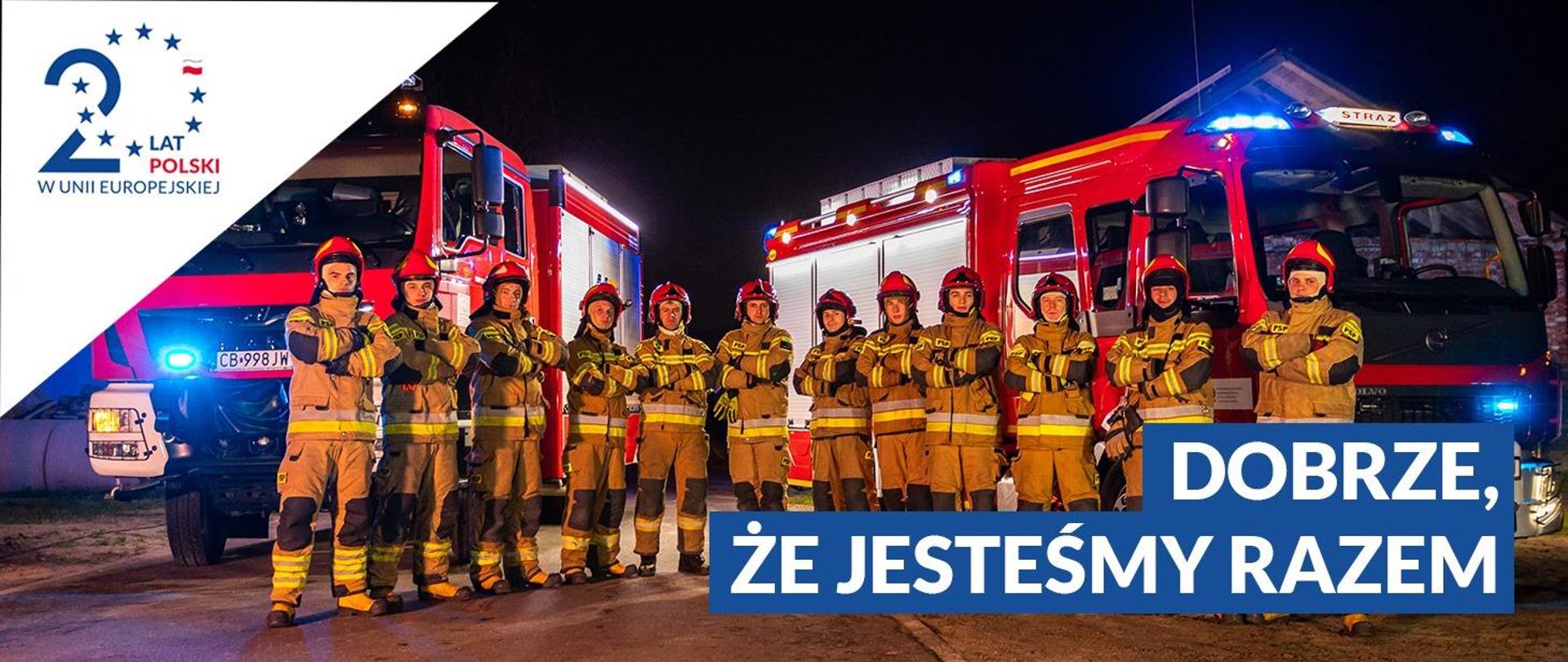 Zdjęcie grupy strażaków stojących pomiędzy dwoma samochodami ratowniczo-gaśniczymi, w prawym dolnym rogu napis Dobrze, że jesteśmy razem. W lewym górnym rogu logotyp 20 lat Polski w Unii Europejskiej