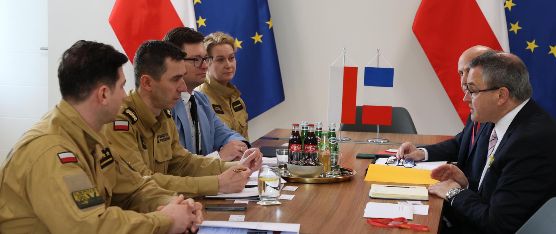 Uczestnicy spotkania siedzą przy stole podczas rozmów. Na stole stoją miniatury flag Polski i Francji. W tle wisi godło polskie oraz stoją na stojakach flagi Polski i Unii Europejskiej.