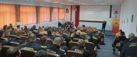 Strażacy siedzą na auli w krzesłach przed nimi stoi mężczyzna z mikrofonem obok stoi baner Komendy Wojewódzkiej PSP w Gdańsku na ekranie wyświetlana jest prezentacja. 