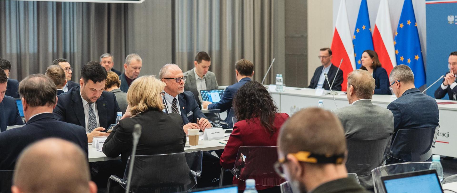 Grupa osób siedzi w sali konferencyjnej przy stołach. W tle flagi PL i UE.