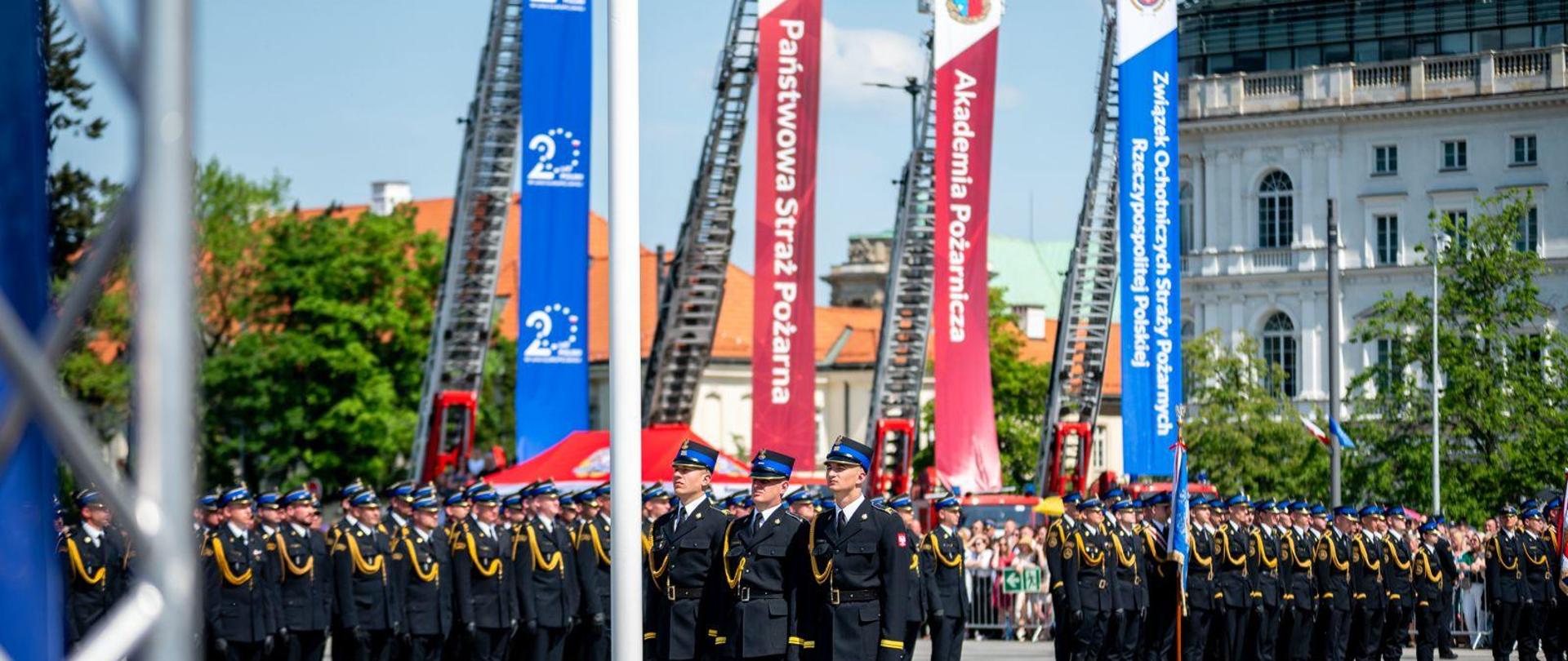 trzech strazaków stoi przy maszcie z flagą, w tle stoją pododdziały strażaków 