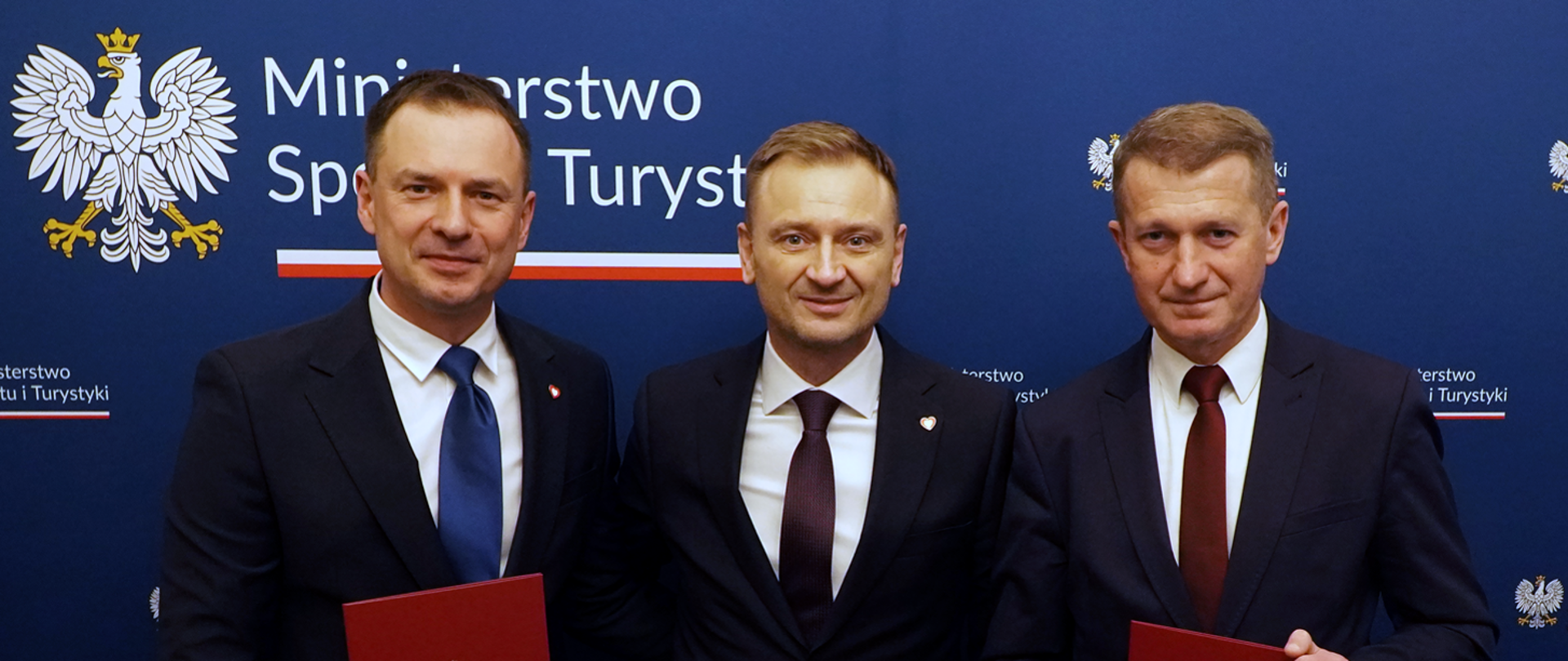 three men in suits standing with the Ministry of Sport and Tourism banner in the background