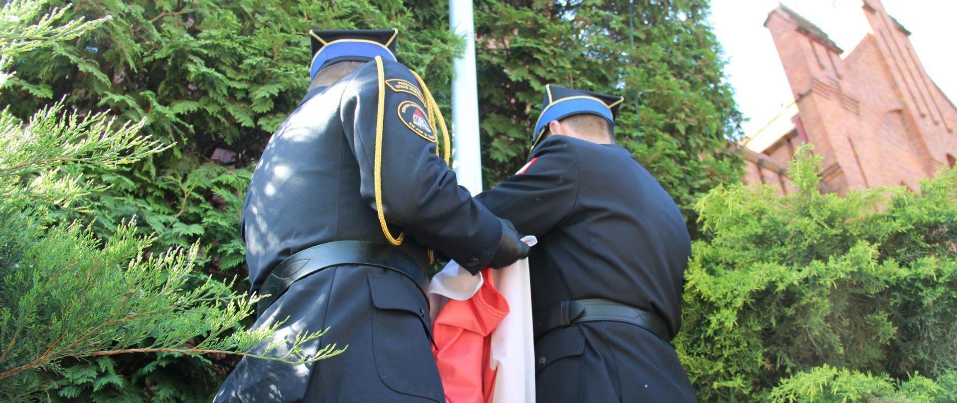 Dwaj oficerowie w mundurach galowych mocują flagę biało-czerwoną do zaczepów na maszcie. Wokół krzewy ozdobne, w dali fragment budynku.