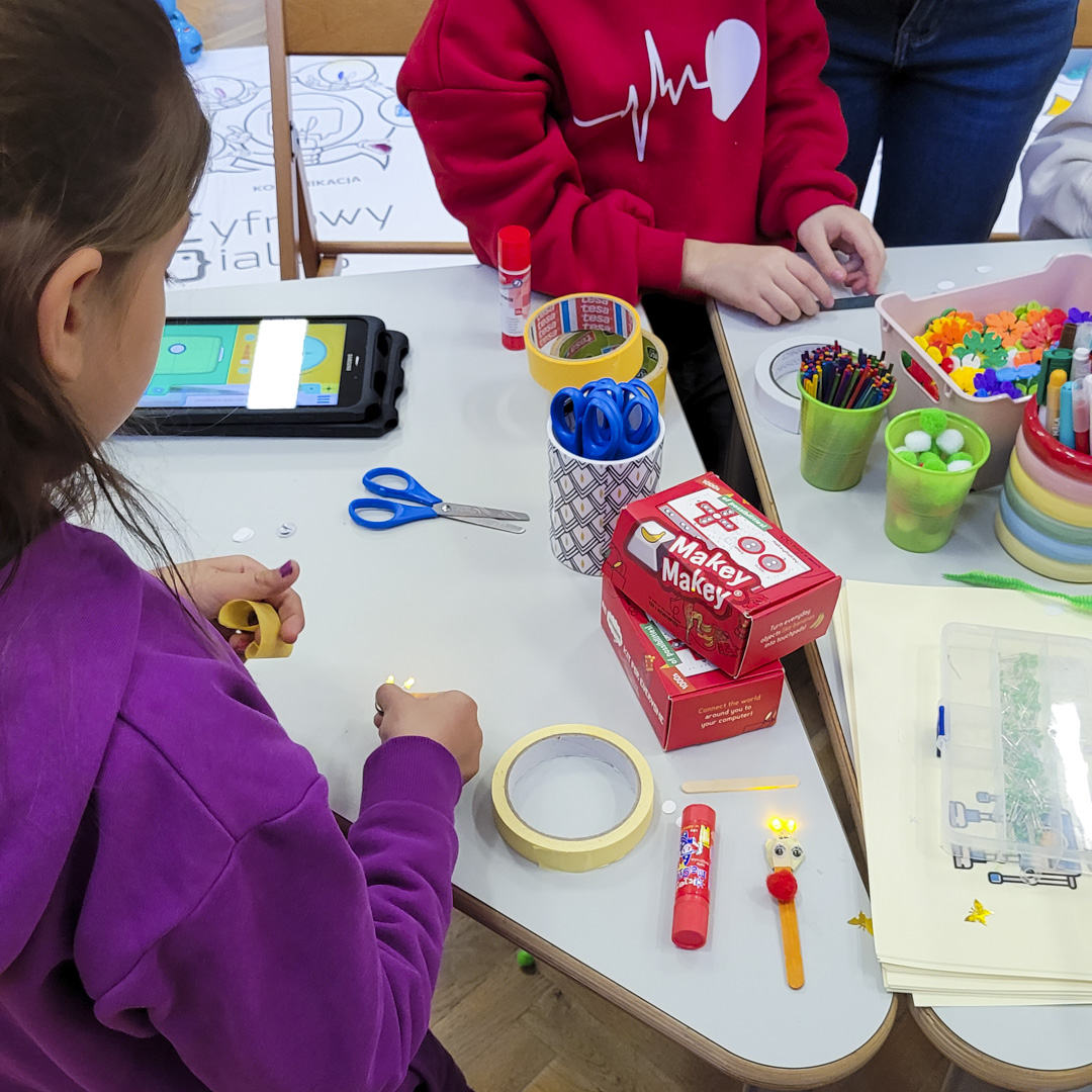 Dzieci przy roboczym stole na którym znajdują się różnorodne materiały podczas warsztatów kreatywnych na CODEWEEK 2024.