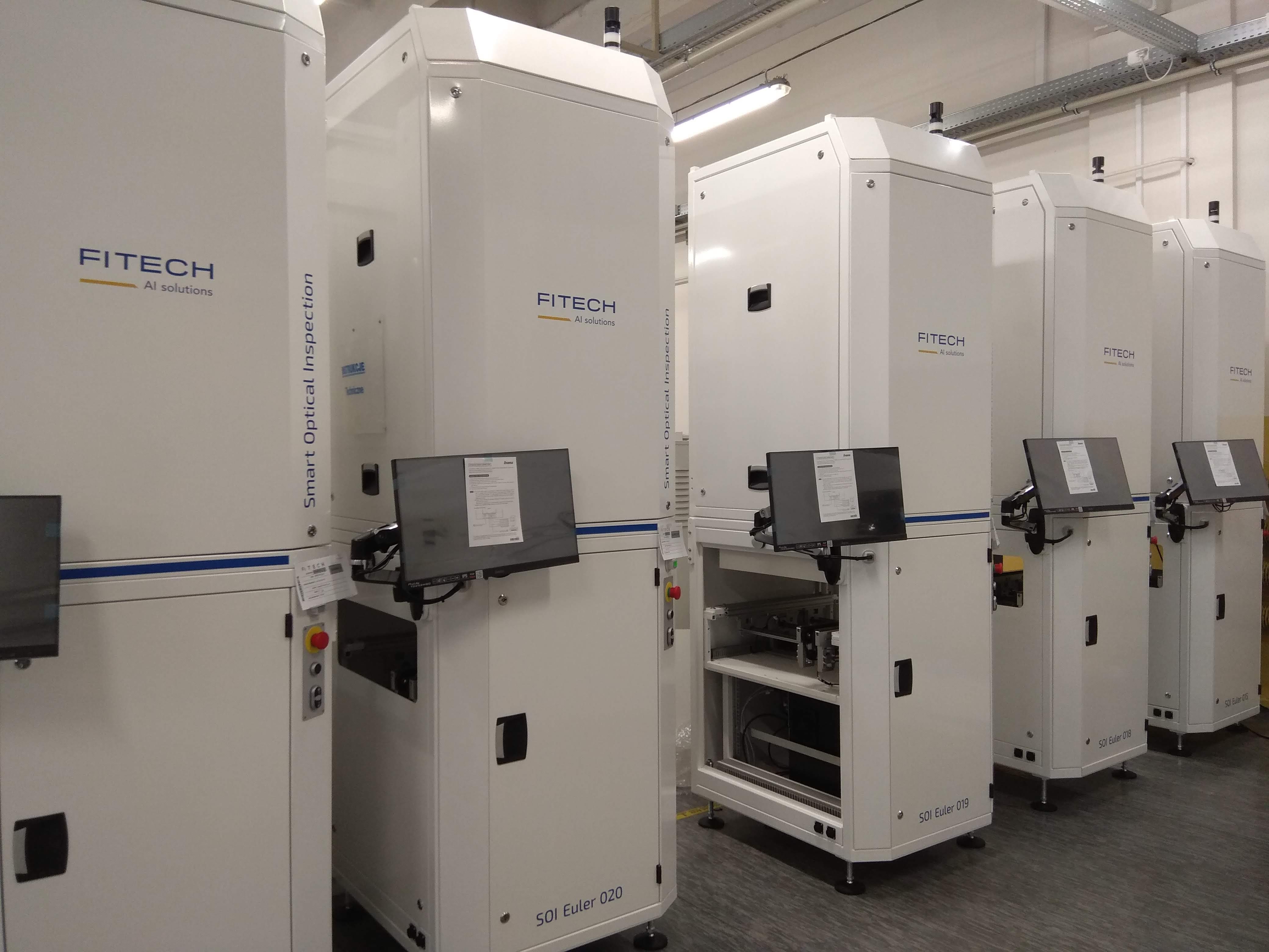 Five ready-made devices, equipped with cuboid monitors in a white housing, stand next to each other in one factory room. On the front, a blue inscription: FITECH AI solutions. Perpendicular to it, the inscription on the side of each device: Smart Optical Inspection. At the bottom of the machines, an inscription: SOI Euler 020.