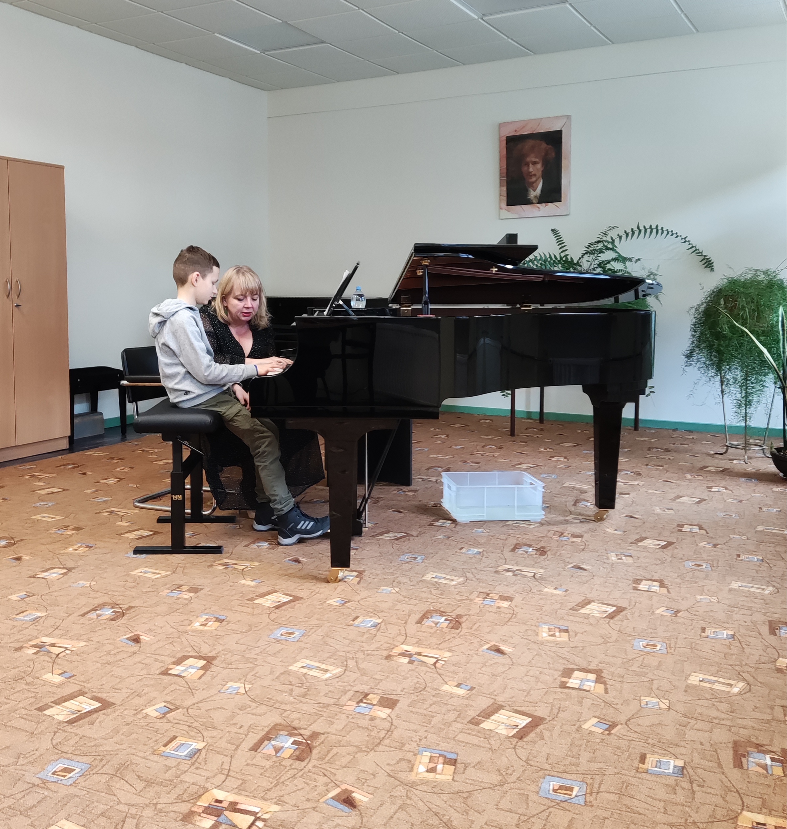 Uczeń siedzący przy fortepianie, obok niego nauczycielka. Po lewej stronie widoczna szafa. Na ścianie wisi portret Ignacego Jana Paderewskiego. Po prawej stronie widać kwiaty.