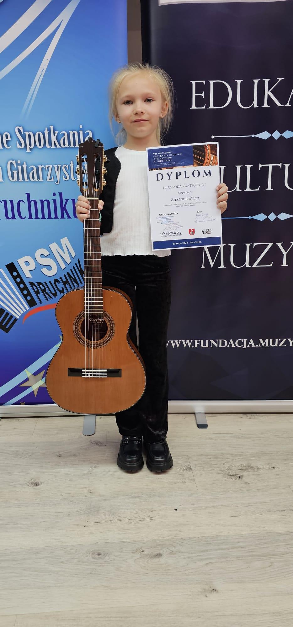 Zuzanna Stach - VII Wiosenne Spotkania Młodych Gitarzystów w Pruchniku. Uczennica z gitarą i dyplomem, w tle reklama konkursu.
