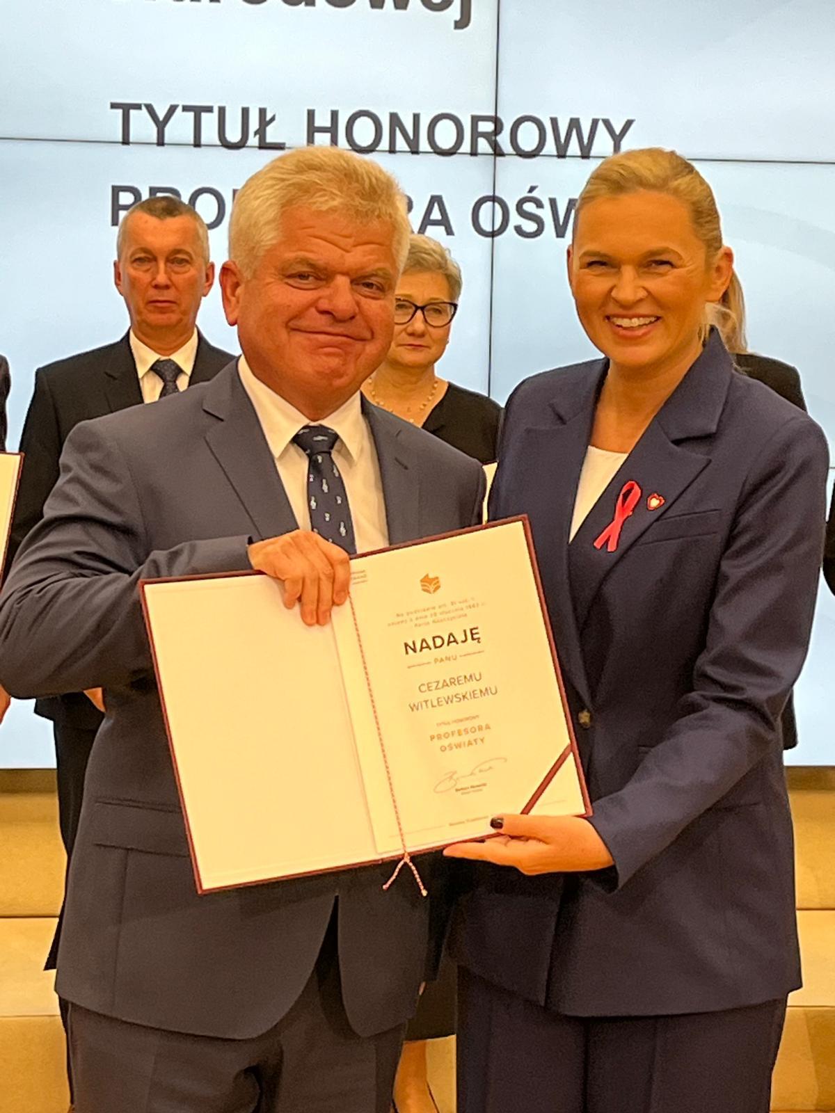 mężczyzna i kobieta ubrani na galowo trzymający przed sobą dyplom, za nimi kilka osób stojących w rzędzie, w tle biały baner