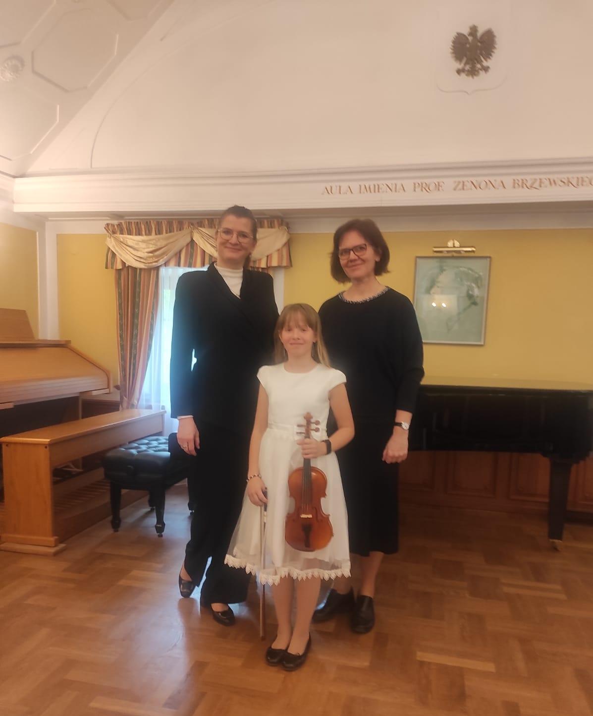 Uczennica w białej sukience trzymająca skrzypce, a po jej bokach nauczycielki. Z tyłu widoczny fortepian oraz organy.