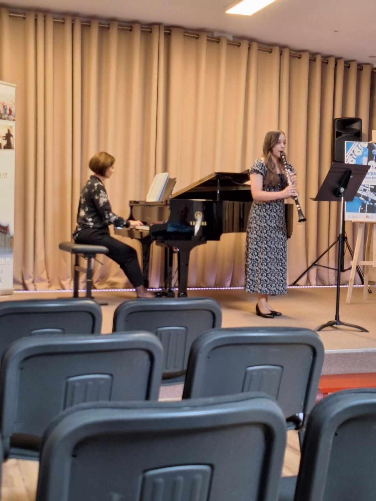 Uczennica grająca na klarnecie oraz towarzysząca jej akompaniatorka na fortepianie. Przed uczennicą widoczny pulpit z nutami. Na dole zdjęcia znajdują się czarne krzesła.