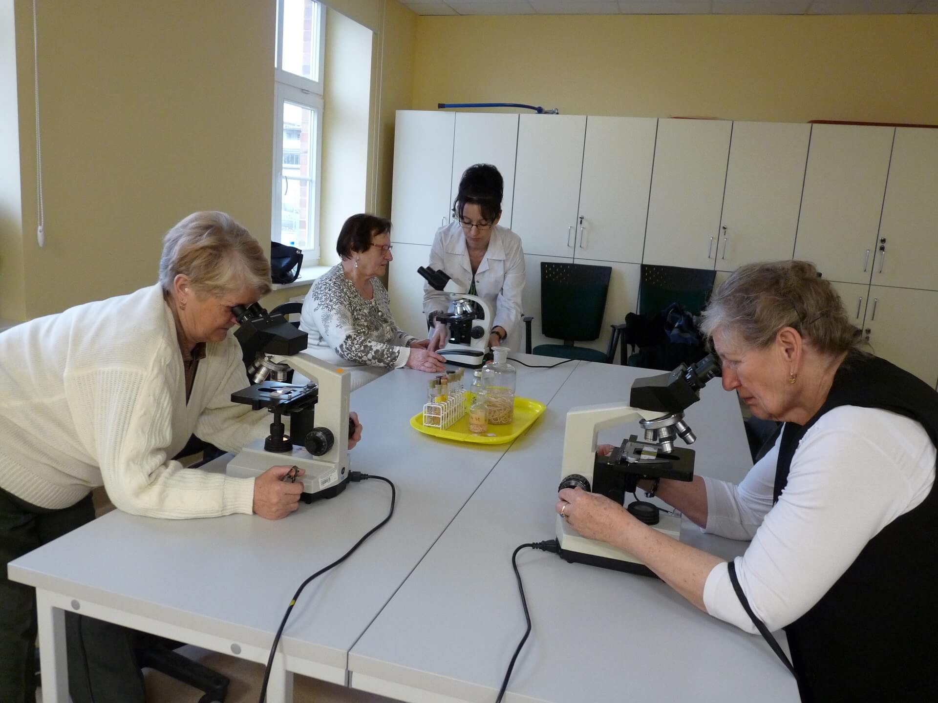 Classes at the Faculty of Biotechnology and Animal Breeding with the participation of a group of seniors. Three ladies are observing microscopes under the watchful eye of a guide wearing a white smock