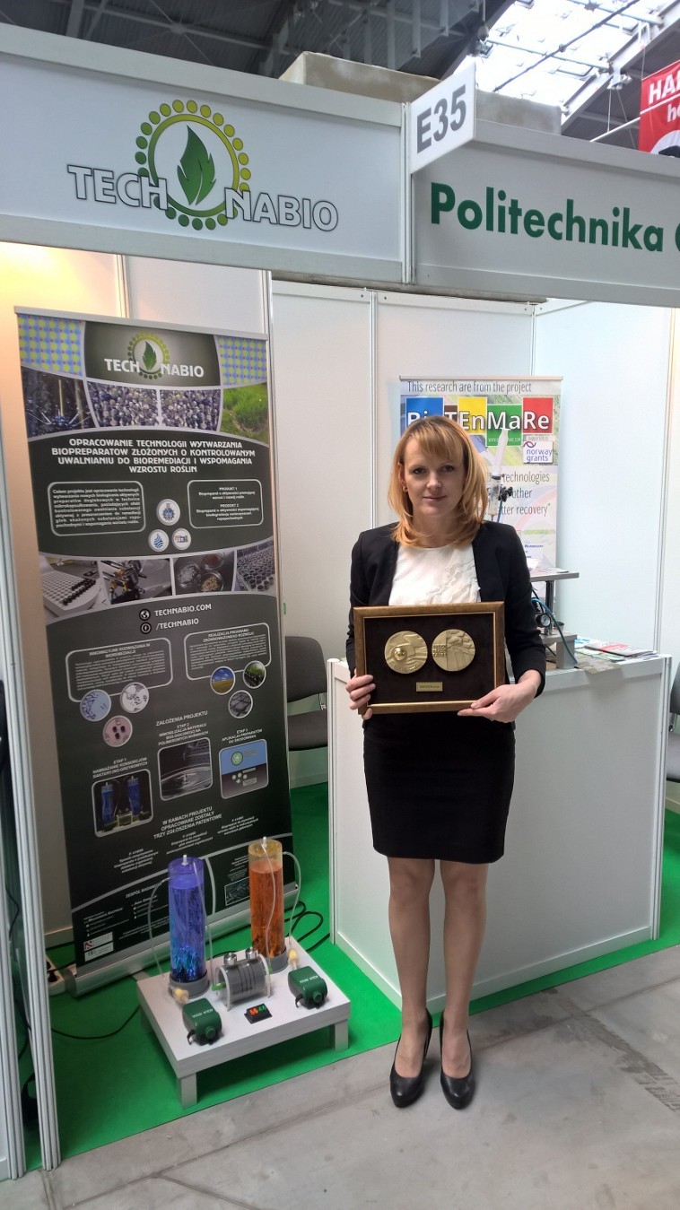 Elegantly dressed dr hab. Anna Grobelak presents the medals which awarded the TechNabio project. In the background, a stand with posters about the project and two measuring cups with colored liquids
