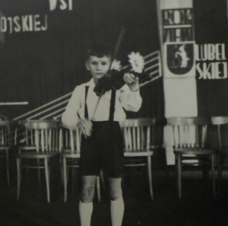 Czarno-białe zdjęcie z lat 1963/1964. Na zdjęciu uczeń Krzysztof Stopa w stroju szkolnym gra na skrzypcach. Z tyłu elementy sceny, krzesła, banery, kotary.