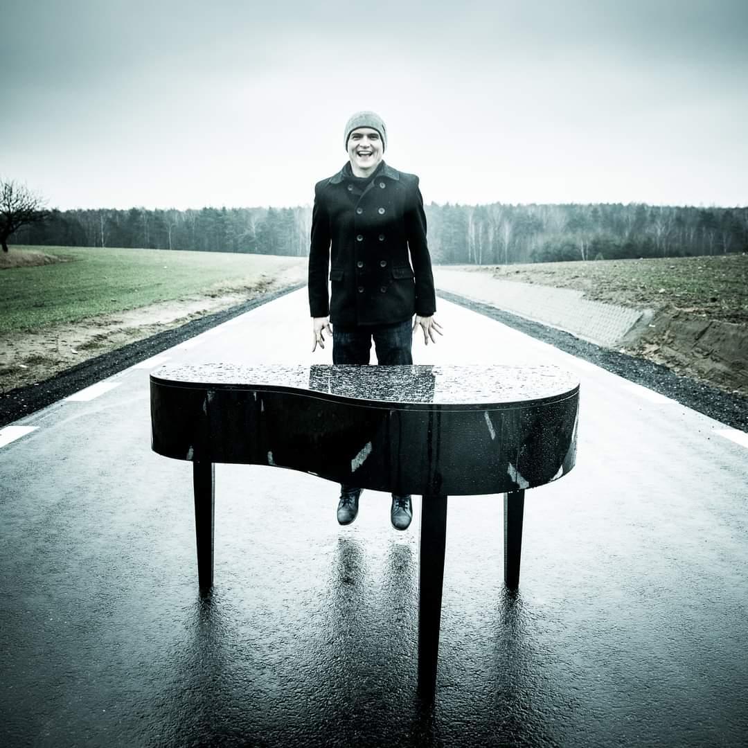 Zdjęcie przedstawia zroszony deszczem czarny fortepian stojący na ulicy pośród pól. Za nim unosi się nad ulicą Maciej Tubis, który ubrany jest w czarny, dwurzędowy krótki trencz i szarą czapkę. W oddali widać las. 