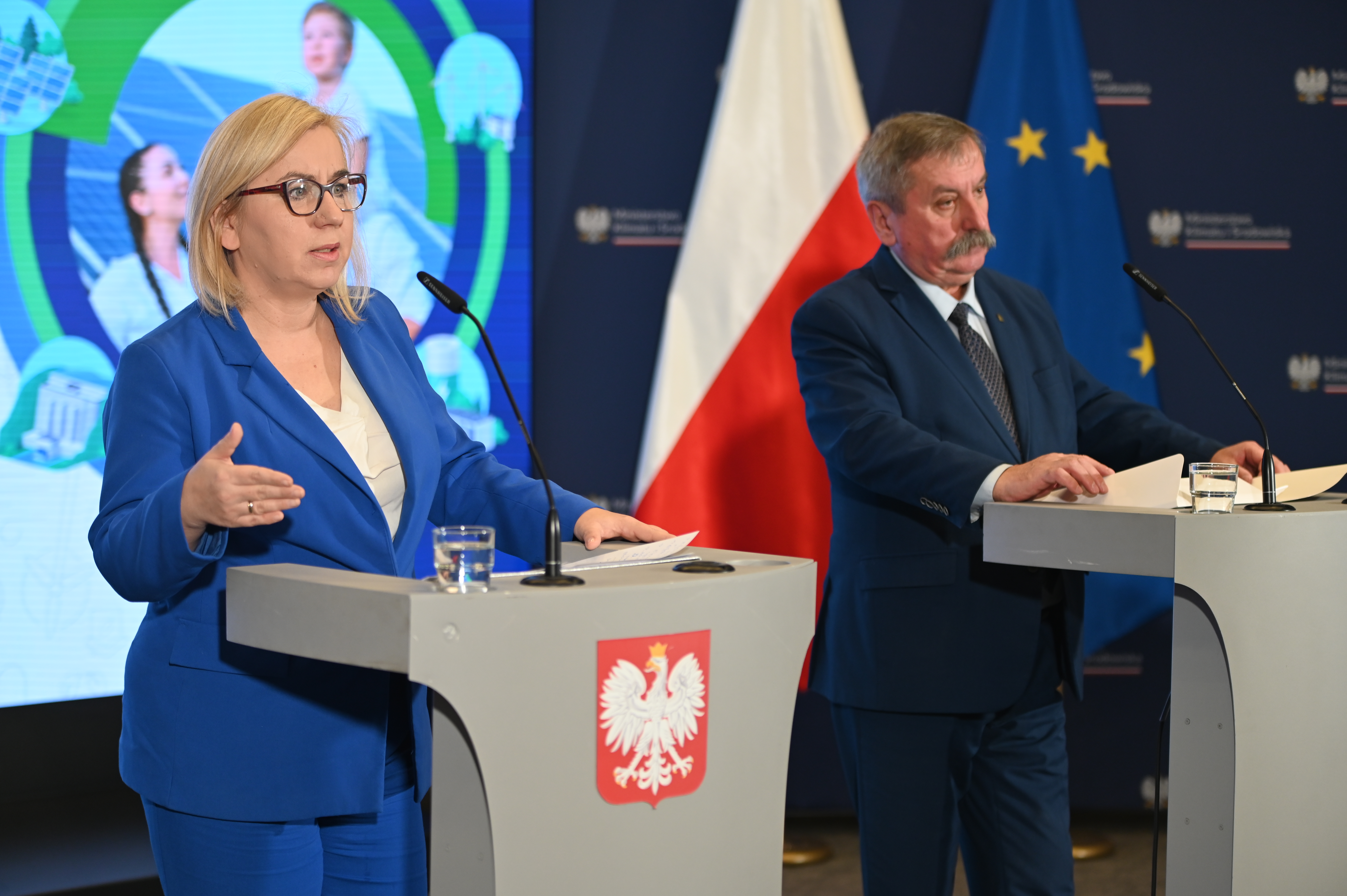 Konferencja ministry klimatu i środowiska Pauliny Hennig-Kloski oraz zastępcy prezesa zarządu NFOŚiGW Józefa Matysiaka, ogłoszenie II naboru wniosków "Energia dla wsi". Przedstawiciele obu instytucji stoją na tle telebimu z informacją o programie "Energia dla wsi" przed mównicami. Widać kamerę nagrywającą ich wystąpienie. 