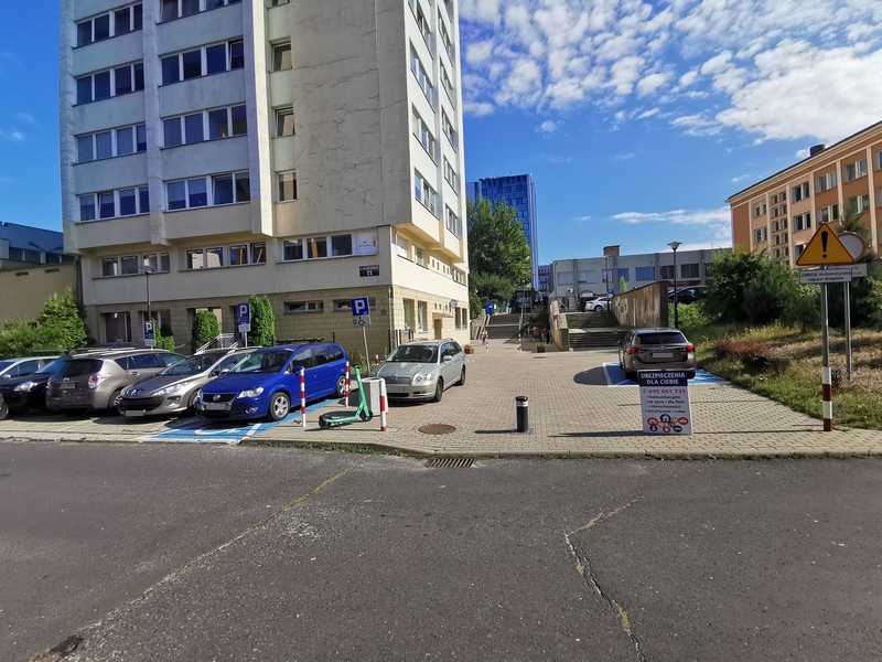 Zdjęcie przedstawia parking z samochodami przy biurowcu, w którym znajduje się Inspektorat, na którym widać dwa oznaczone miejsca do parkowania dla osób z niepełnosprawnościami.