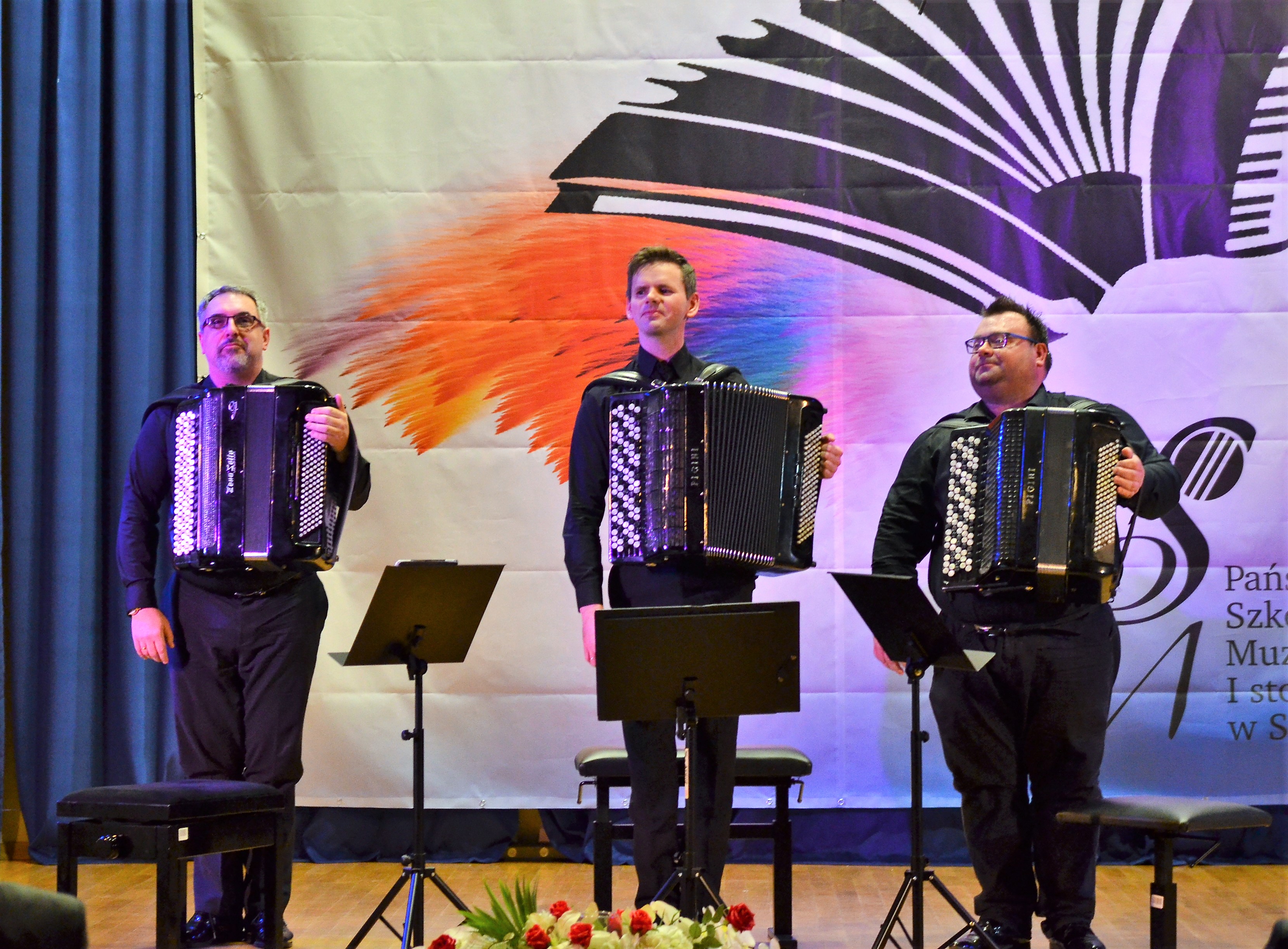 Trio akordeonowe stojące na scenie, za nimi baner konkursu akordeonowego i niebieska kotara