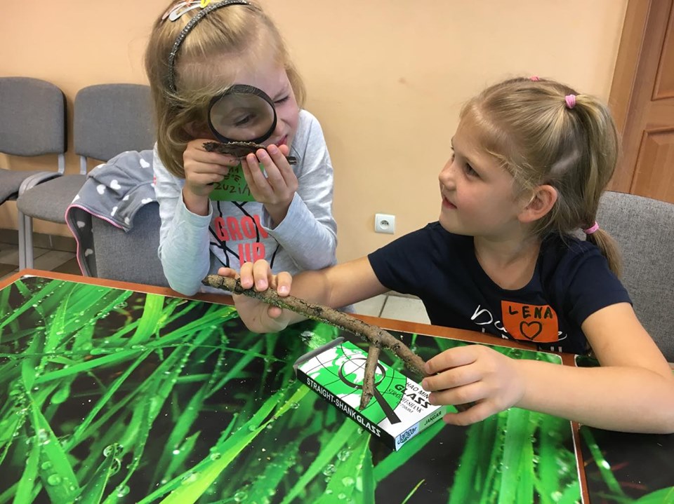 Two about 7-year-old girls at a table covered with a floral pattern. The girl on the left is examining the bark of wood under a magnifying glass. The second girl is holding a dry branch in her hands