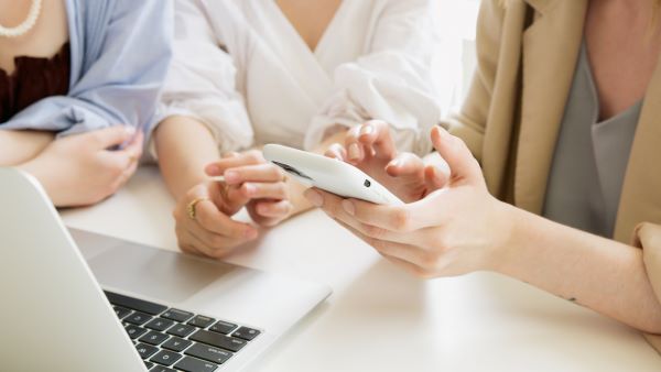 Zdjęcie przedstawiające ręce trzech kobiet siedzących przed laptopem jedna z nich uzywa telefonu komórkowego