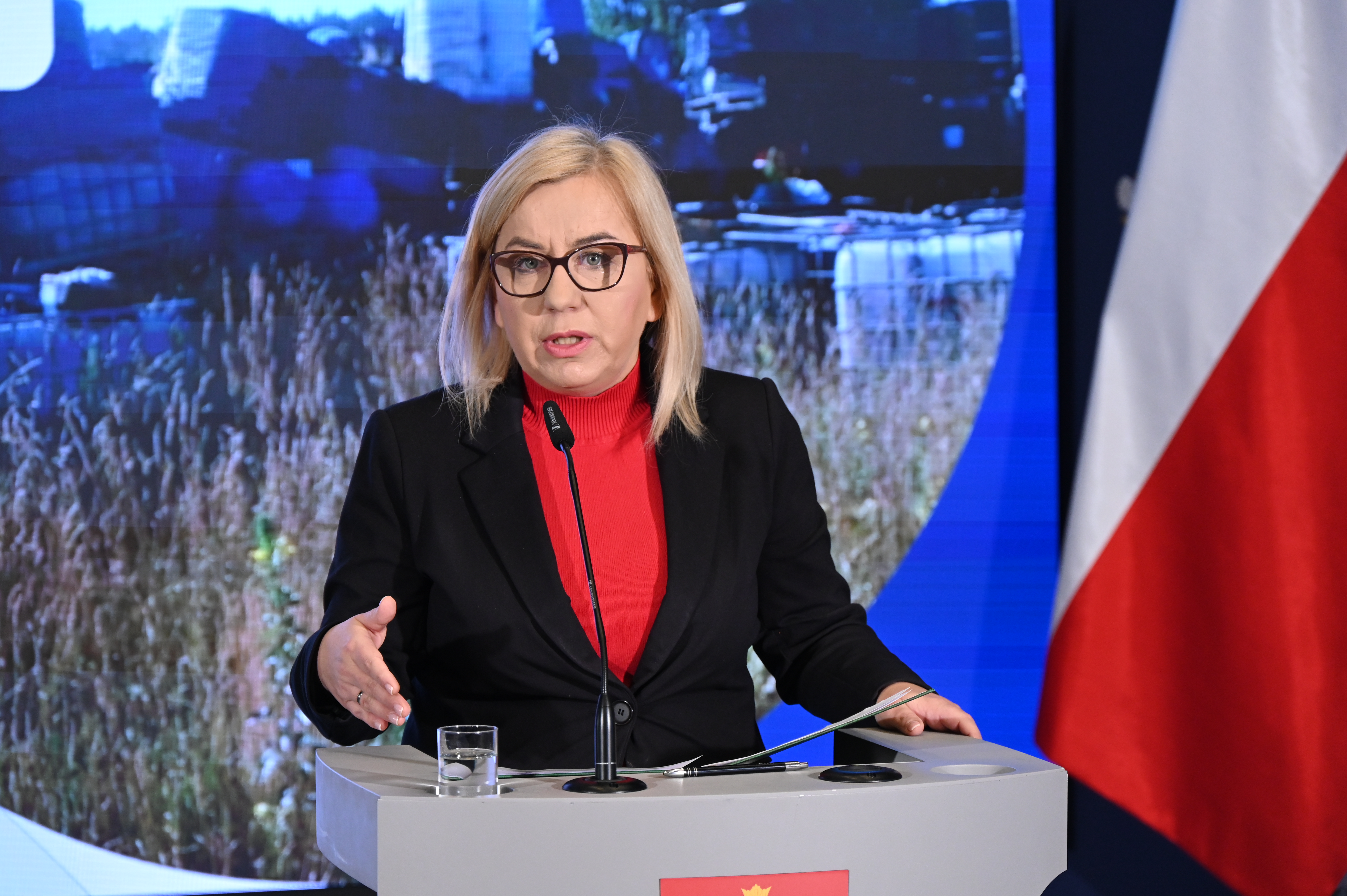 Ministra klimatu i środowiska podczas konferencji prasowej nt. dofinansowania w wysokości 200 mln zł dla samorządów na usuwanie nielegalnie zgromadzonych odpadów. Ministra stoi i mówi do mikrofonu, za nią znajduje się ekran z prezentacją i flaga Polski.