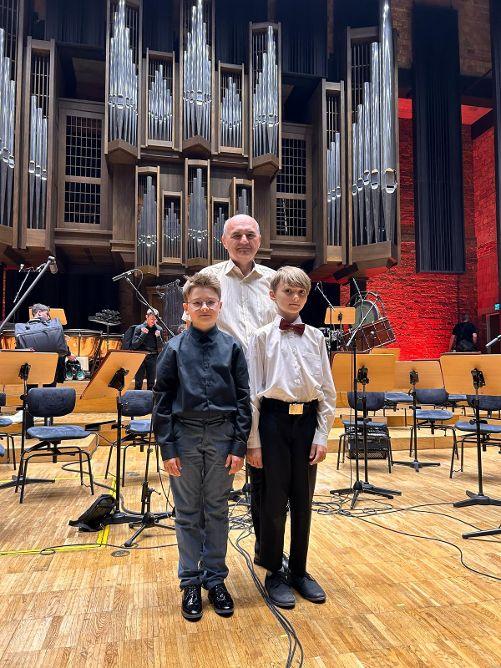 Dwóch uczniów stojących na scenie Filharmonii, a za nimi nauczyciel. Z tyłu widoczne krzesła, pulpity na nuty oraz organy.