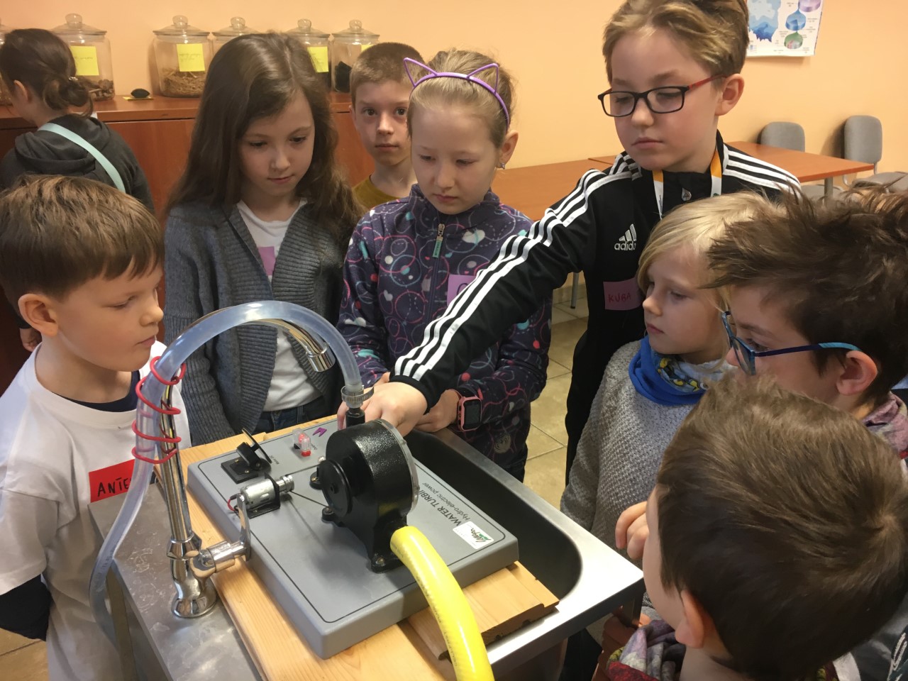 Research laboratory. A group of eight children aged around 8-10 stand around a specialized device. The tallest boy is trying to turn it on