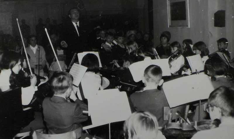 Czarno-białe zdjęcie przedstawiające uczniów z instrumentami smyczkowymi w trakcie gry. Przed uczniami stoi dyrygent. W głębi słuchająca publicznośc.