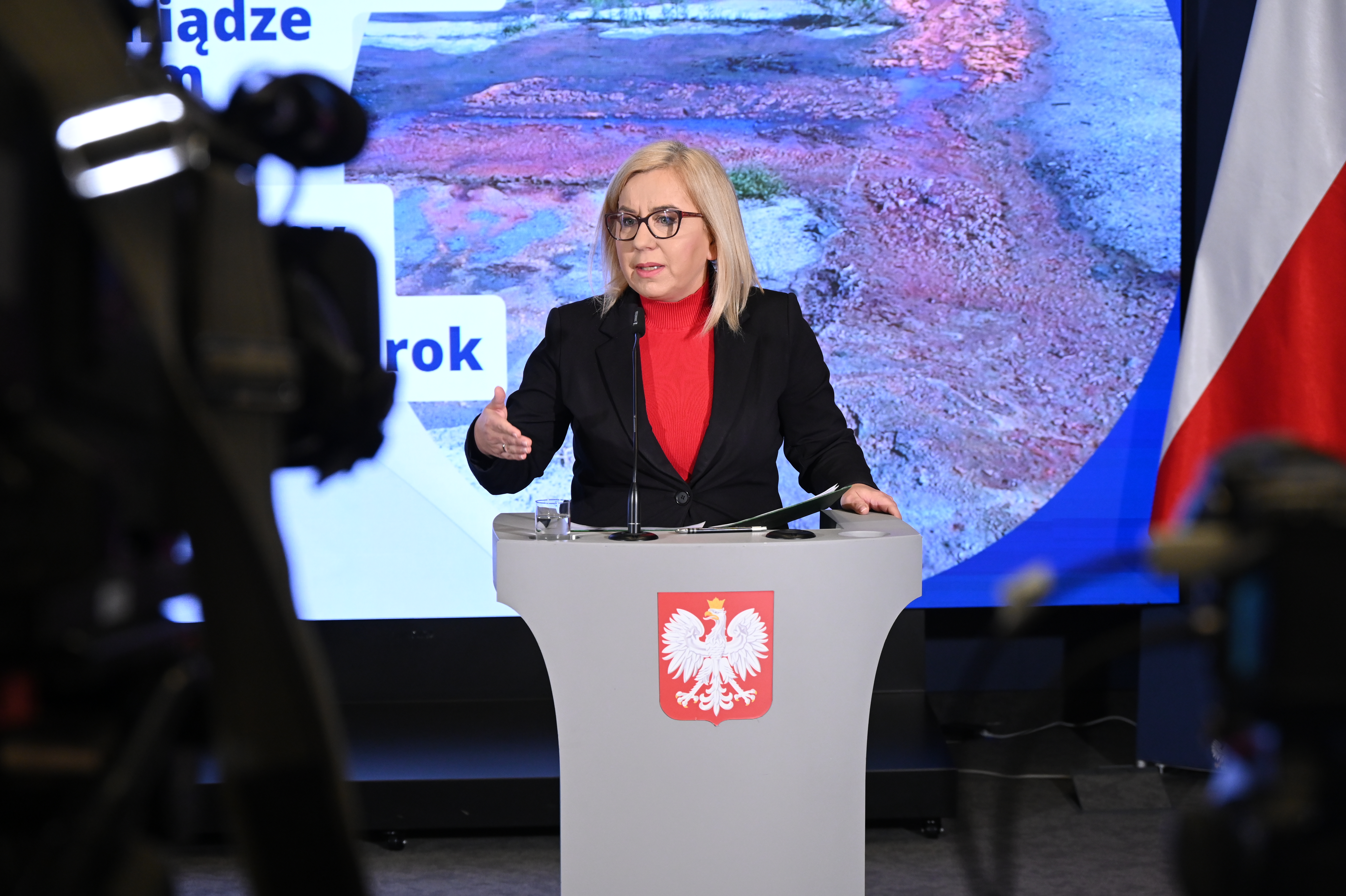 Ministra klimatu i środowiska podczas konferencji prasowej nt. dofinansowania w wysokości 200 mln zł dla samorządów na usuwanie nielegalnie zgromadzonych odpadów. Ministra stoi przy mównicy i mówi do mikrofonu, na pierwszym planie zdjęcia znajdują się kamery.