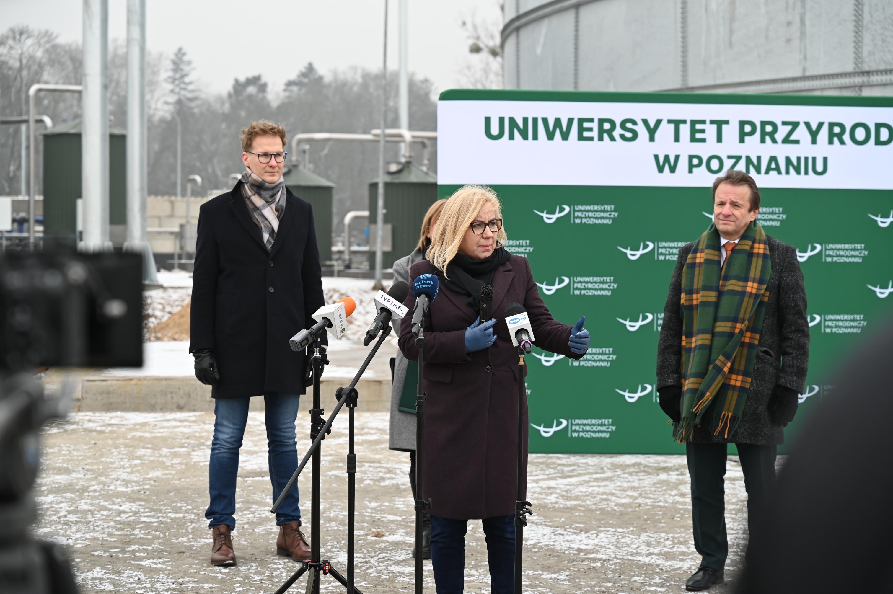 Ministra klimatu i środowiska stoi przed kamerami na konferencji prasowej na terenie Rolniczego Gospodarstwa Doświadczalnego w Brodach. Za nią stoi pozostali prelegenci. Konferencja na tle budynku pierwszej w Polsce biometanowni.
