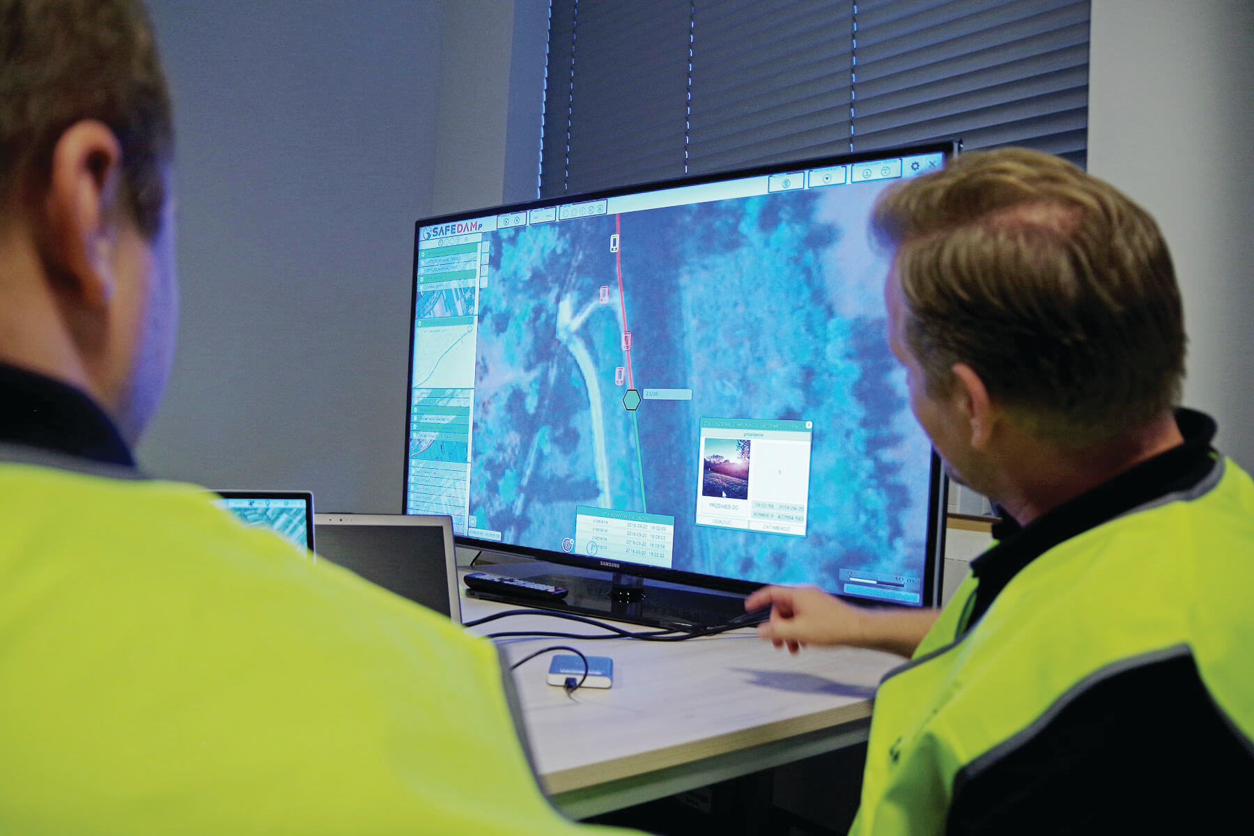 Two men in yellow reflective vests follow the data from the SAFEDAM system on a computer monitor