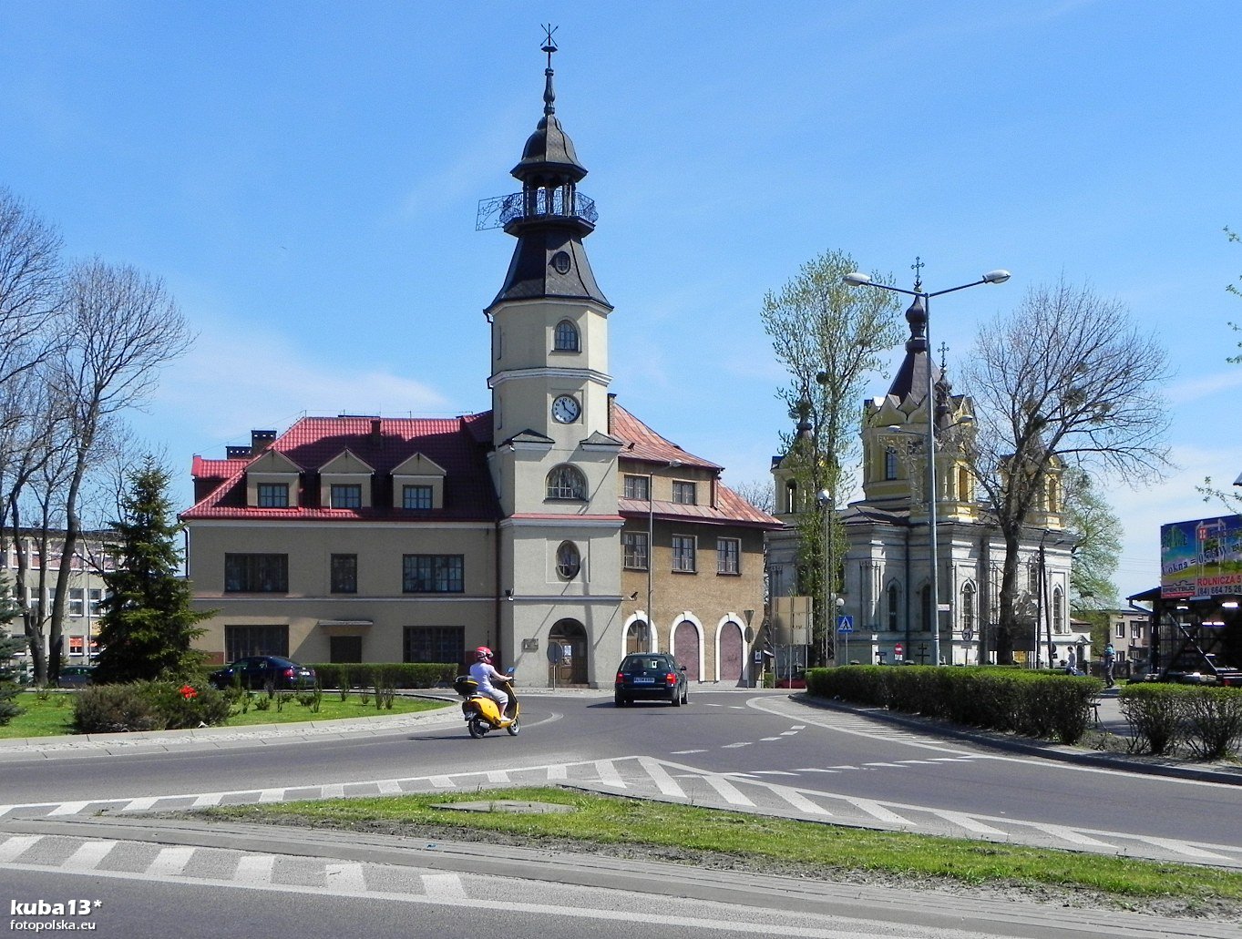 zdjęcie przedstawia budynek PSM od ulicy-ronda, z pojazdami poruszającymi się na rondzie, po bokach drzewa, krzewy, w tle od lewej część budynku biblioteki, od prawej cerkiew prawosławna, na samej górze bezchmurne niebo 