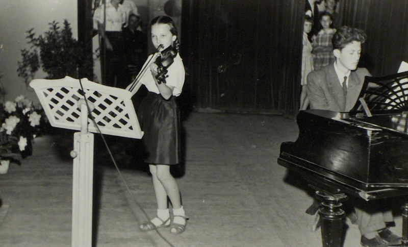 Czarno-białe zdjęcie z 1950 roku, przedstawiające uczennicę Marię Domina w trakcie występu na scenie. Uczennica gra na skrzypcach. Przed nią stoi duży, drewniany pulpit na nuty. Po prawej stronie akompaniator przy fortepianie. Z tyłu kotara zza której wyglądają uczniowie i nauczyciele obserwujący występ zza sceny.