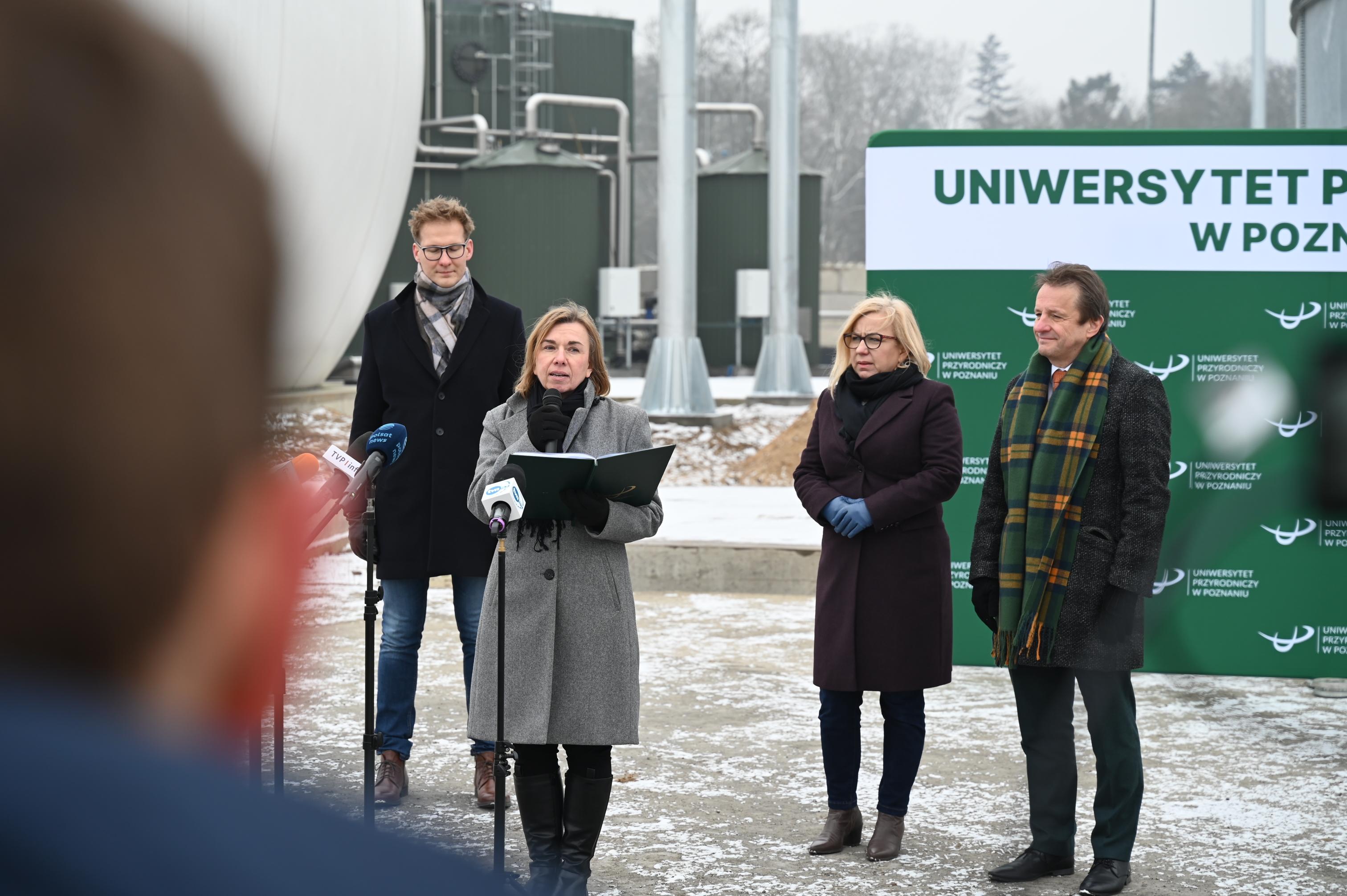 Przemawia rzeczniczka prasowa Uniwersytetu Przyrodniczego Iwona Cieślik. Na dworze, na tle poprzednich rozmówców, przed banerem Uniwersytetu Przyrodniczego w Poznaniu i na tle budynku biometanowni. 