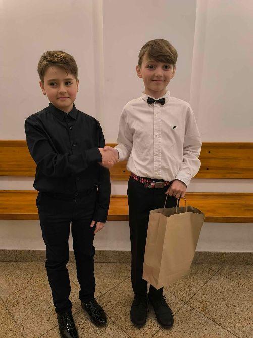 Dwóch uczniów podaje sobie dłoń. Jeden z nich, stojący po prawej stronie trzyma papierową torbę.