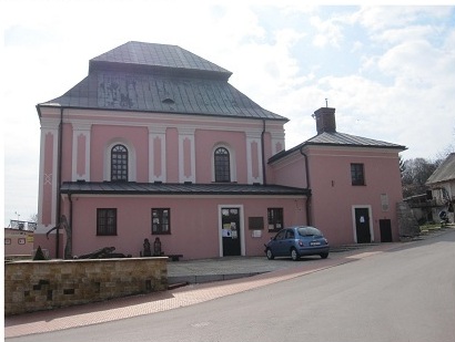 Zdjęcie przedstawia synagogę w Szczebrzeszynie. Budynek w jednopiętrowy z kolorze ciemno pomarańczowym, w tle niebo jasno niebieskie, przed szkołą stoi niebieski samochód