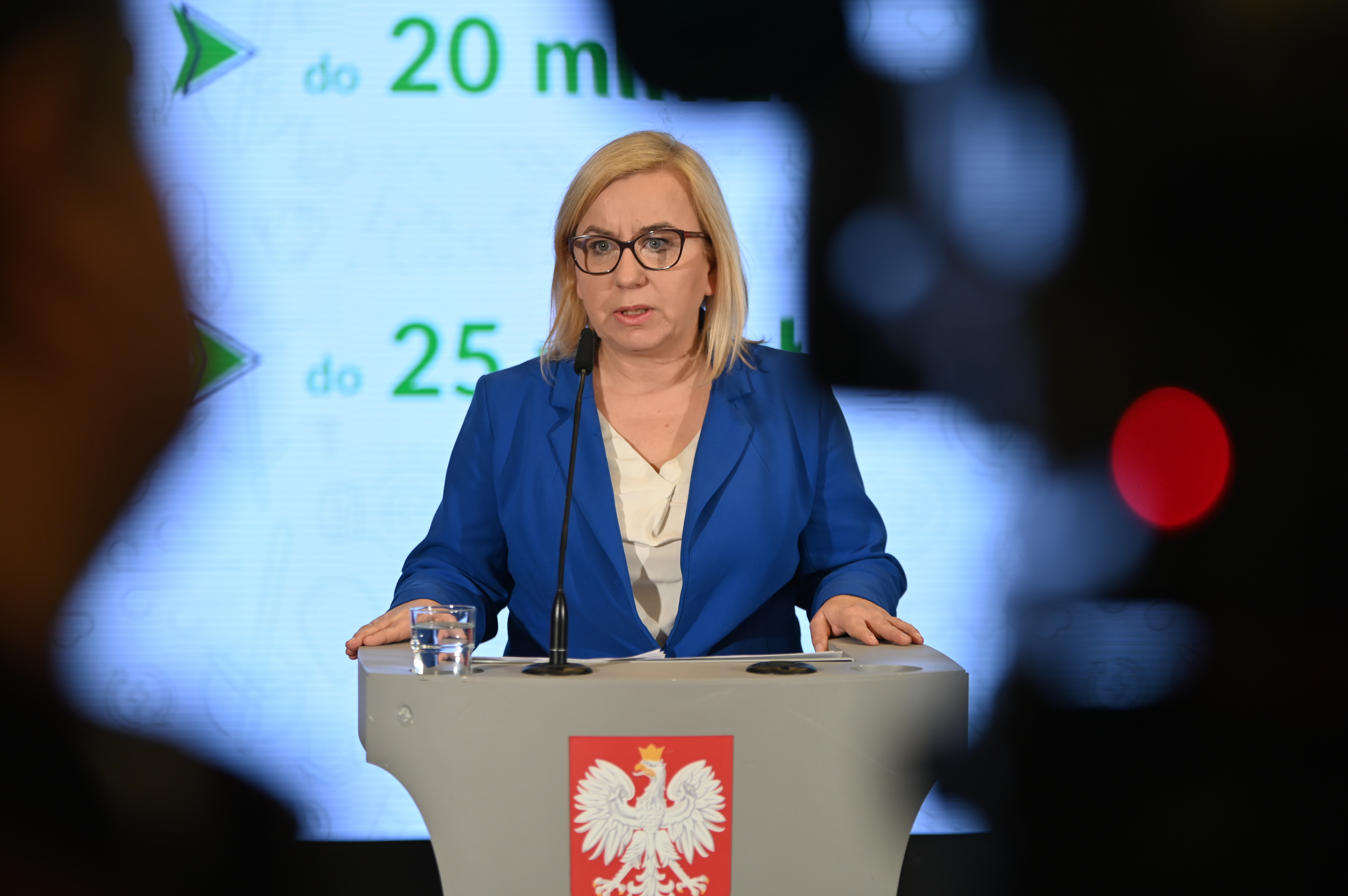 Ministra klimatu i środowiska Paulina Hennig-Kloska stoi za mównicą z godłem Polski i ogłasza na konferencji dla mediów II nabór wniosków do programu "Energia dla wsi". Widać zarys kamer telewizyjnych na tle których wypowiada się ministra.