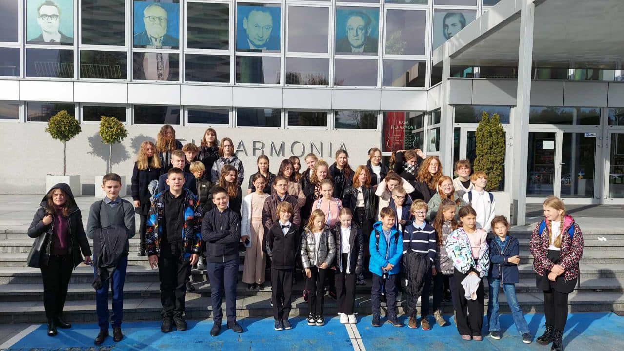 Grupa dzieci wraz z opiekunami stojąca na schodach przed Filharmonią w Rzeszowie. Z tyłu widoczne okna Filharmonii wraz z portretami kompozytorów.