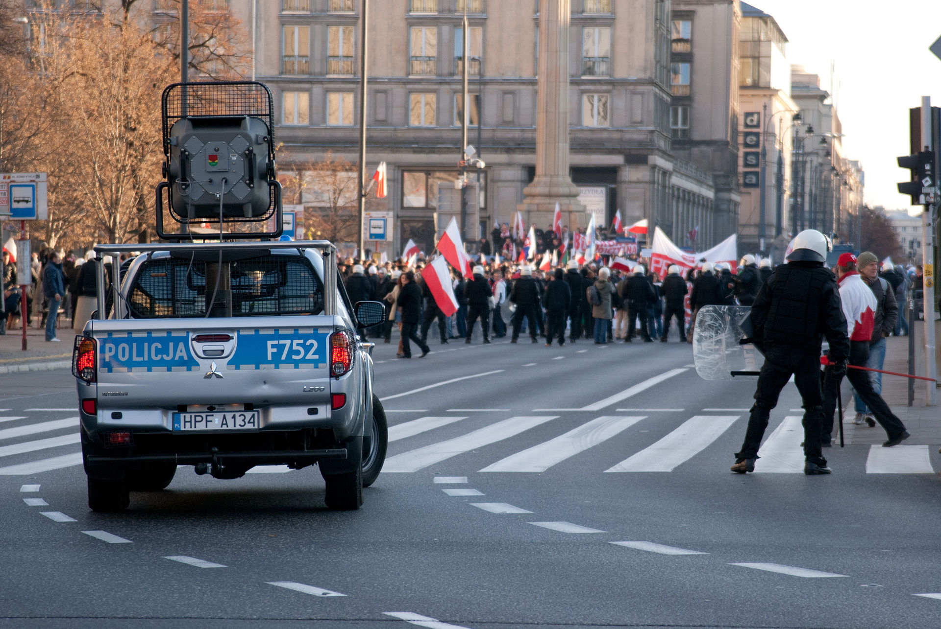 Zdjęcie przedstawia radiowóz Policji oraz maszerujących ludzi z flagami Polski.