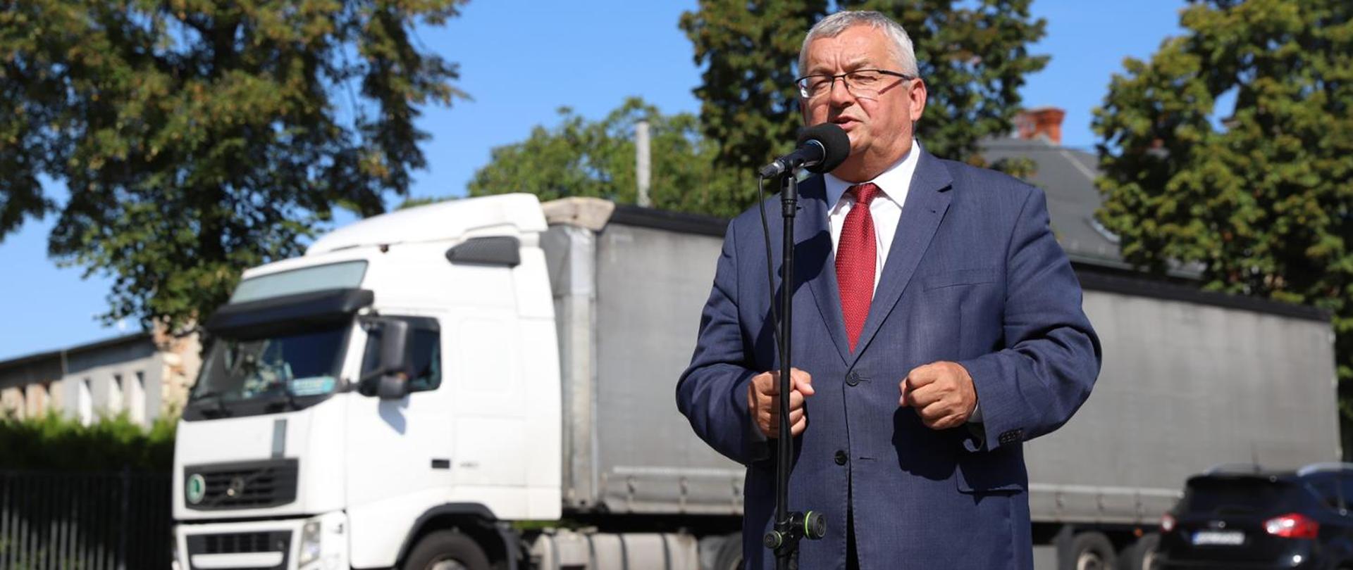 Minister infrastruktury Andrzej Adamczyk wypowiadający się podczas konferencji prasowej dotyczącej budowy obwodnicy Wschowy. W tle przejeżdżający przez miejscowość samochód ciężarowy.