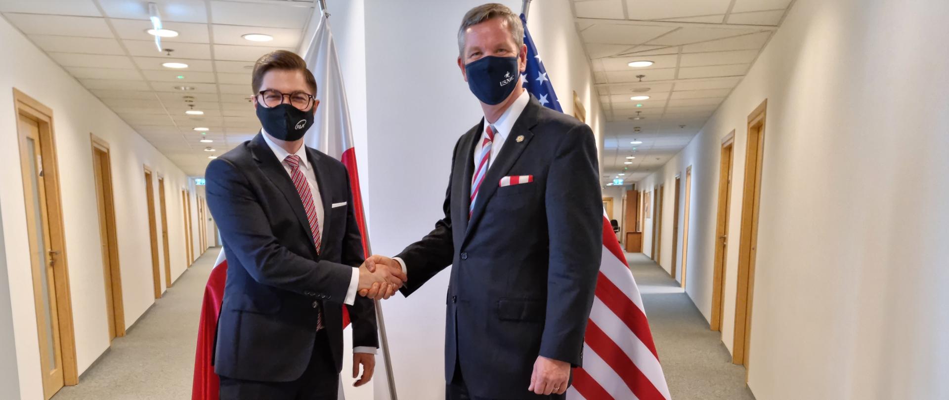 Meeting of the President of the National Atomic Energy Agency with the Chairman of the Nuclear Regulatory Commission. Dr. Łukasz Młynarkiewicz and Chistopher T. Hanson are shaking hands. In the background, the Polish and American flags.