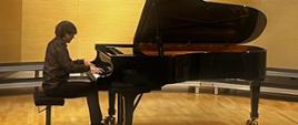 Zdjęcie przedstawia artystę grającego na fortepianie na scenie w sali koncertowej szkoły. Fortepian czarny marki Fazioli. Mężczyzna ubrany na czarno.