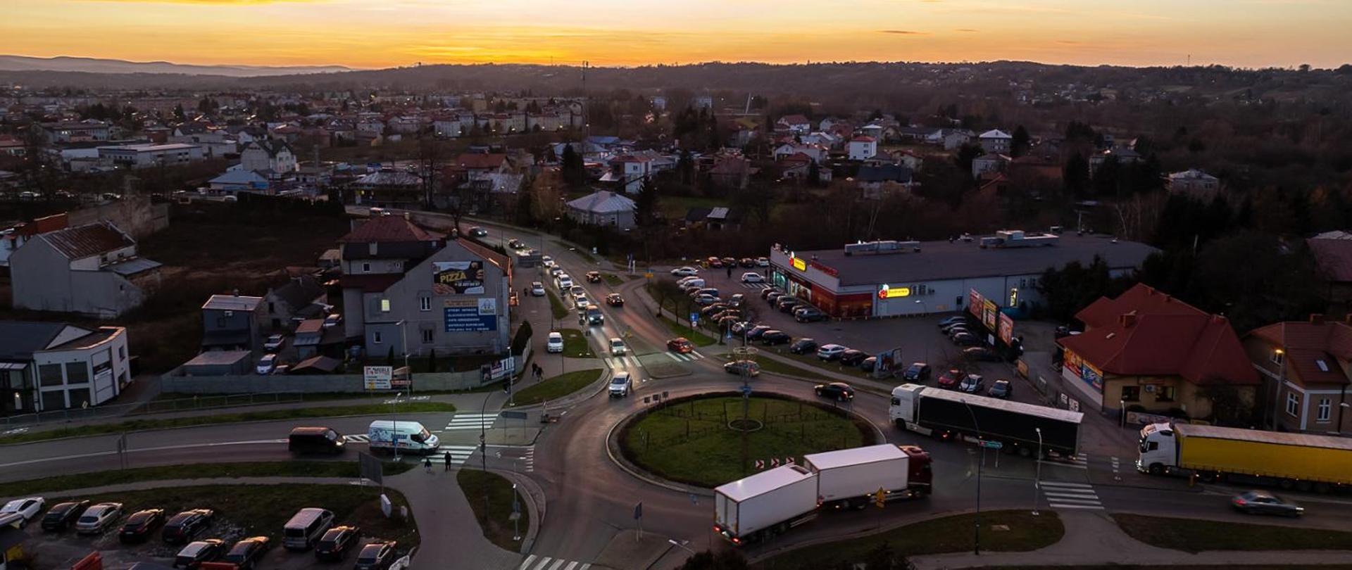 Widok z góry na rondo na DK73 w Jaśle. Widać auta stojące w korku na każdym wlocie na rondo. Są to auta osobowe i ciężarowe. Pora popołudniowa, widać powoli zmierzch. 