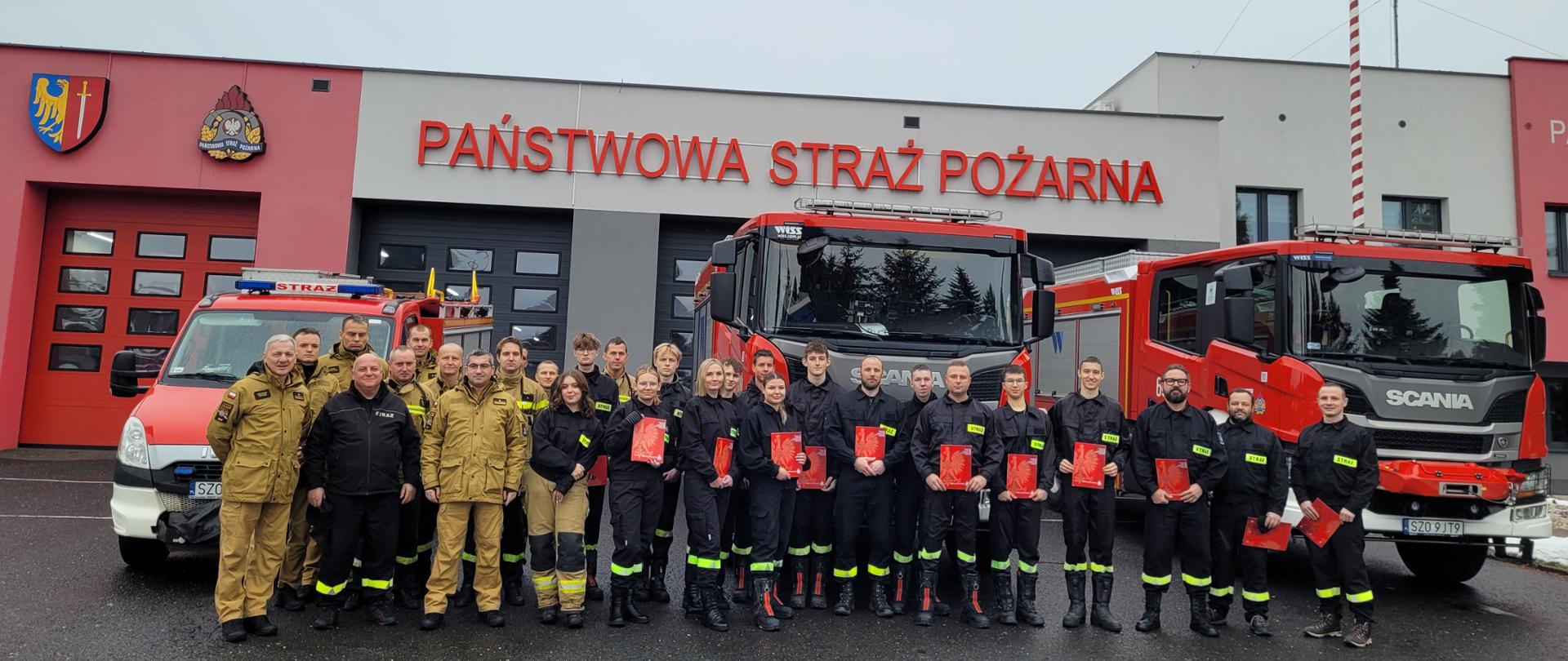 Strażacy stoją na tle trzech samochodów pożarniczych ustawionych na tle budynku garażu. Kursanci trzymają w rękach czerwone teczki. 